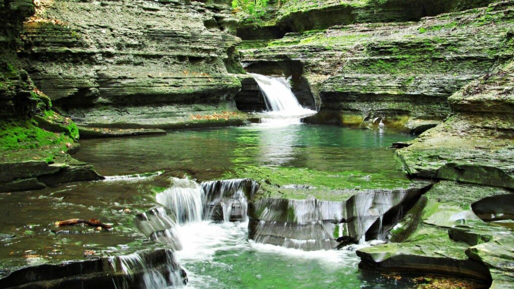 Cascading waterfall over rocky cliffs with lush green grass and clear water pool landscape wallpaper