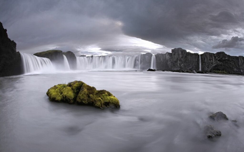 Wide waterfall over rocky cliffs with mossy stone in reflective water under cloudy sky wallpaper