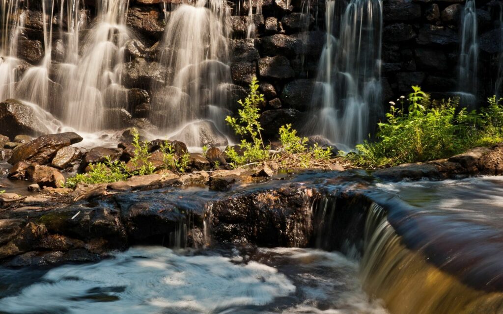 Tranquil waterfall over dark rocks with white foam and lush green riverbank in natural light Wallpaper