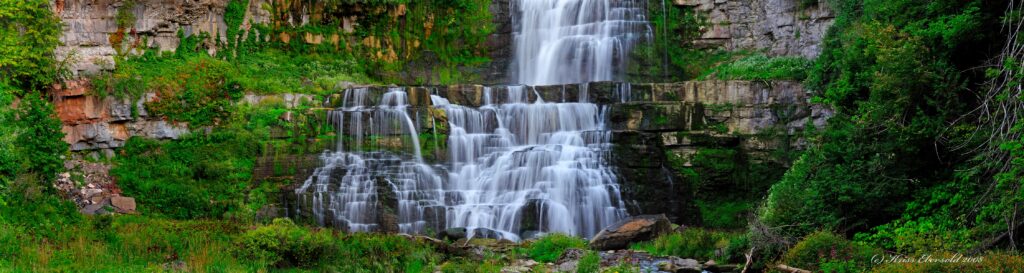 Waterfall cascading over layered rocks into lush green landscape with mossy stones and foliage Wallpaper