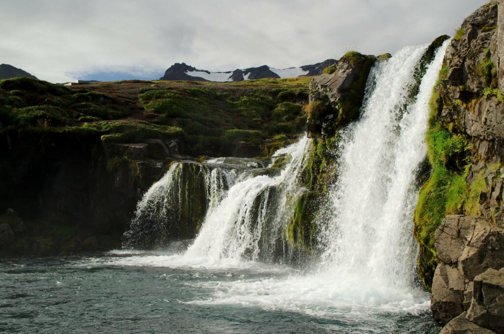 Cascading waterfall over mossy rocks into a clear river with rugged mountains under cloudy sky Wallpaper