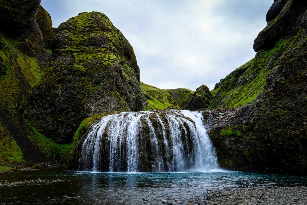 Waterfall cascading over mossy rocks into clear blue river with cliffs and green hills background Wallpaper