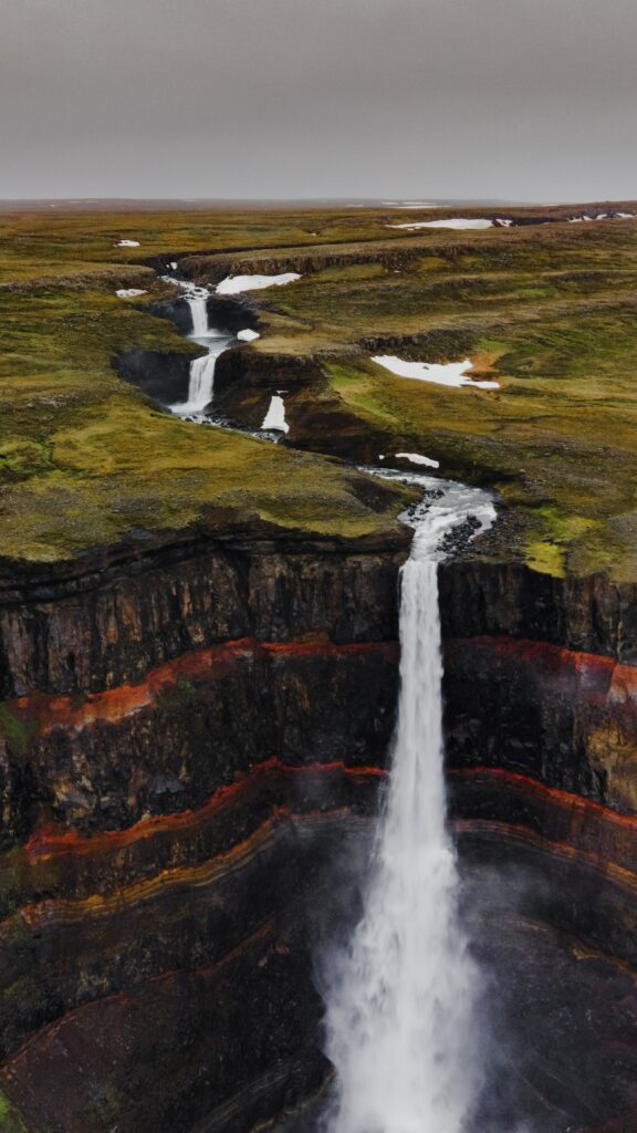 Waterfall cascading over rugged cliffs with winding stream and lush green grass background Wallpaper