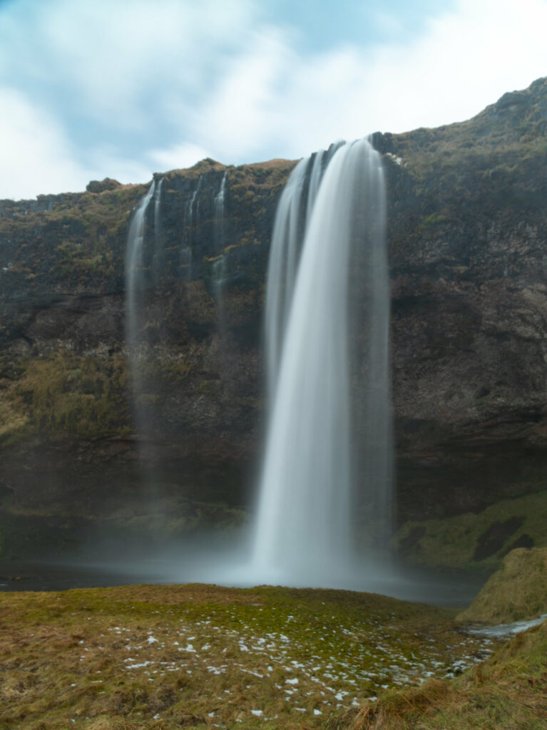 Waterfall cascading over rugged cliffs with green grass and snow in a serene natural setting Wallpaper