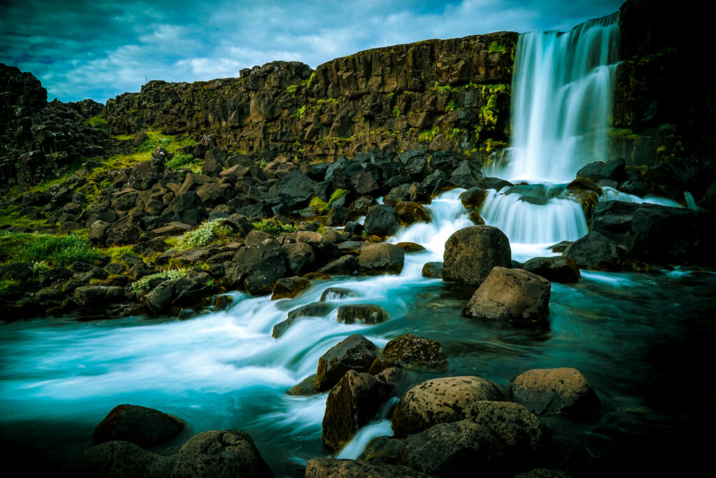 Tranquil waterfall flowing over rocks into clear blue pool surrounded by lush green grass and stones Wallpaper