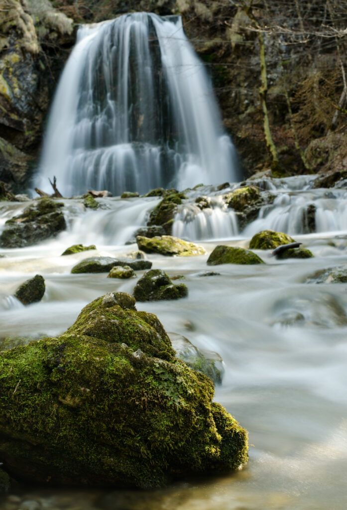 Tranquil waterfall flowing over rocky stones into river surrounded by lush moss and greenery Wallpaper