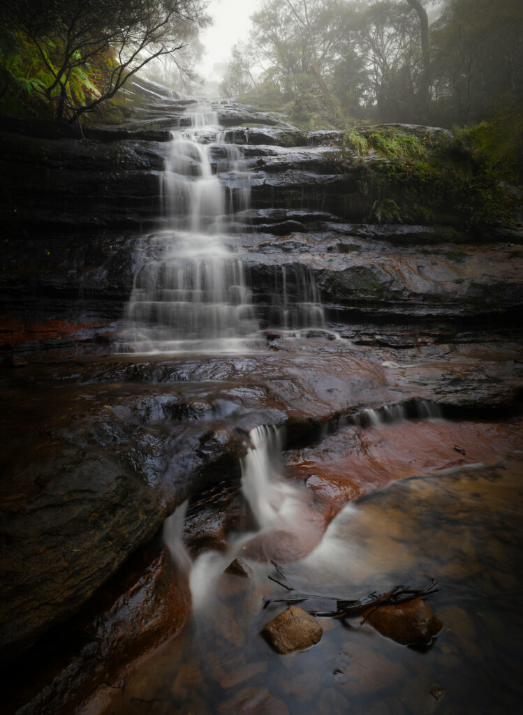 Serene waterfall cascading over smooth stones with lush greenery and misty forest background Wallpaper