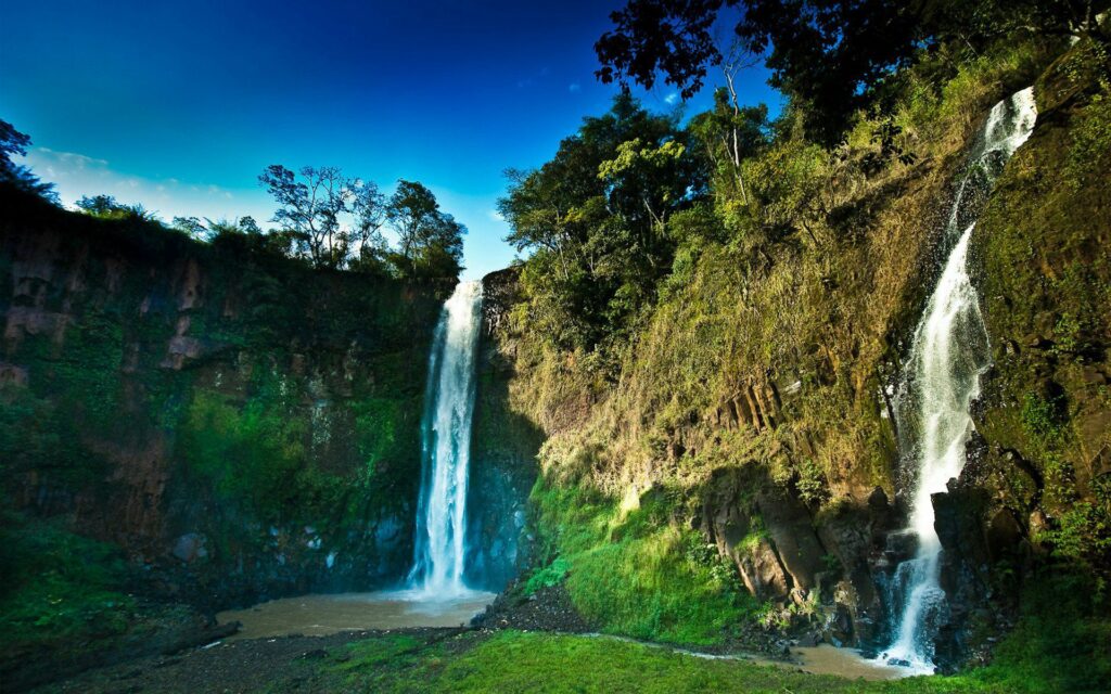 Vibrant waterfall cascading over rocks with lush moss and tall trees under clear blue sky wallpaper