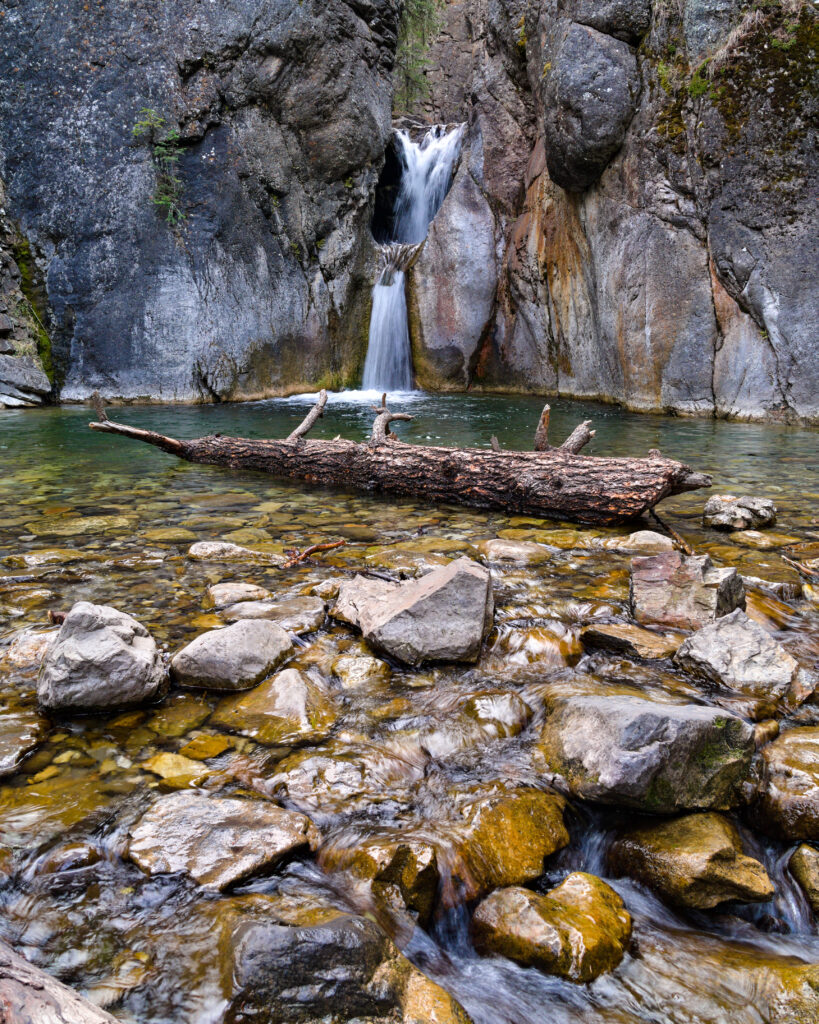Tranquil waterfall flowing over rugged rocks into clear pool with stones and fallen log forest Wallpaper