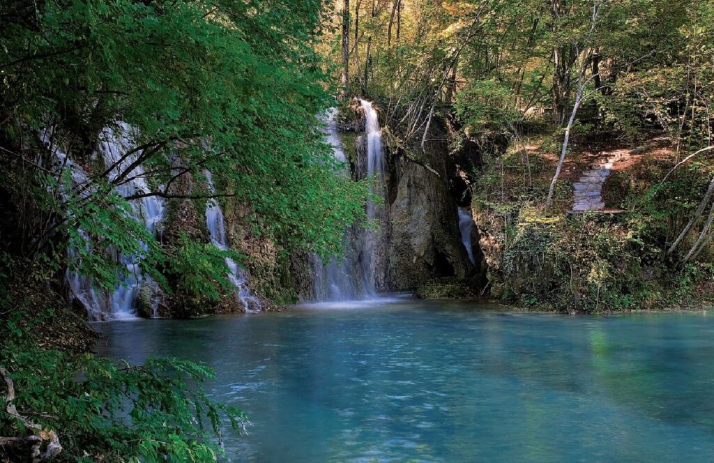 Tranquil waterfall flowing into blue pool surrounded by lush green trees and natural rocks wallpaper