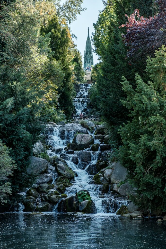 Tranquil waterfall over smooth stones by lush forest with distant Gothic cathedral under blue sky Wallpaper