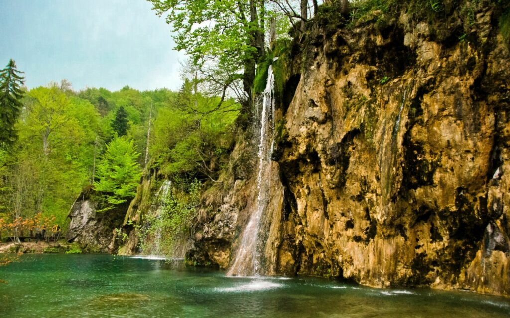 Tranquil river flowing beneath waterfall surrounded by lush green forest and mossy stones Wallpaper