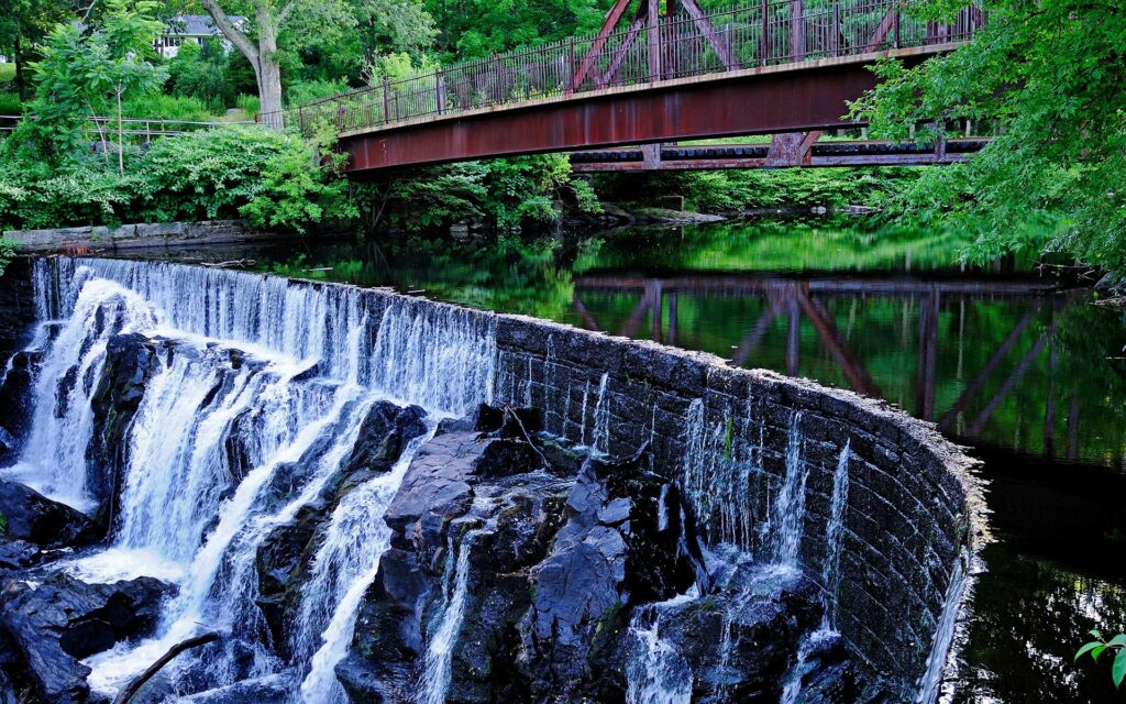 Waterfall cascading over rocks by tranquil river with lush greenery and rustic iron bridge background Wallpaper