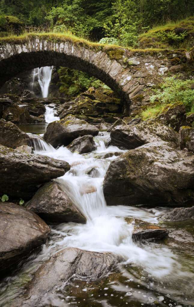 Serene waterfall cascading over stones under stone bridge surrounded by lush green nature background Wallpaper