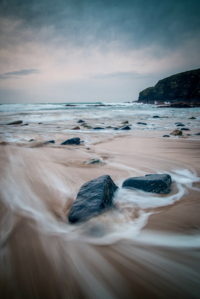 Dynamic ocean waves swirling around smooth shore rocks under pastel skies and distant cliffs Wallpaper