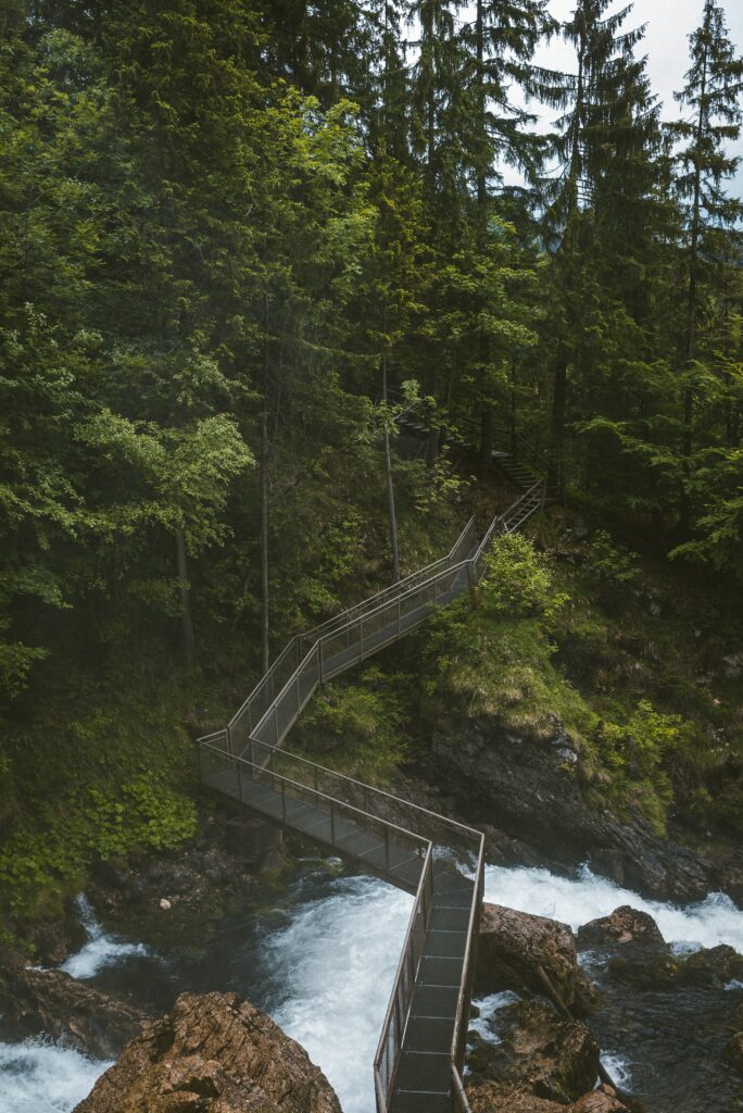 Winding wooden bridge over rushing river with mist and dense green forest landscape background Wallpaper