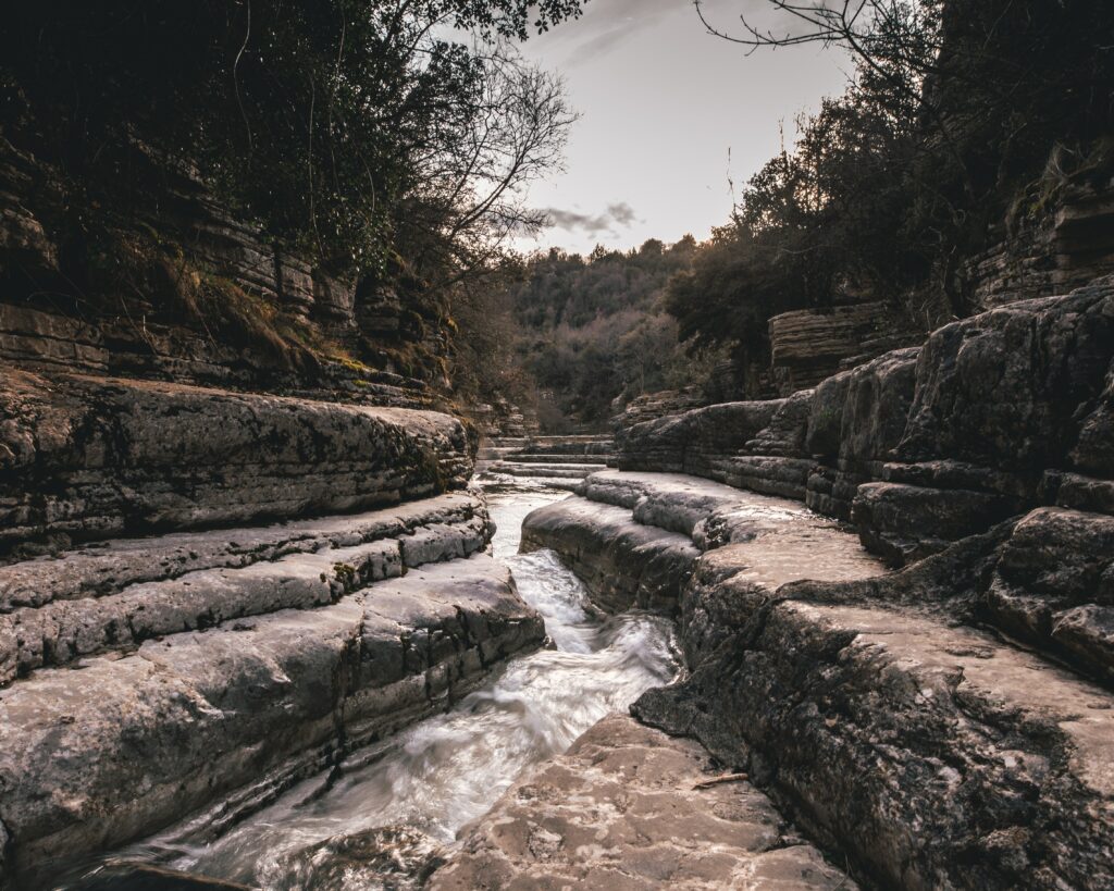 Winding stream flowing through rocky terrain with leafless trees under overcast moody sky wallpaper