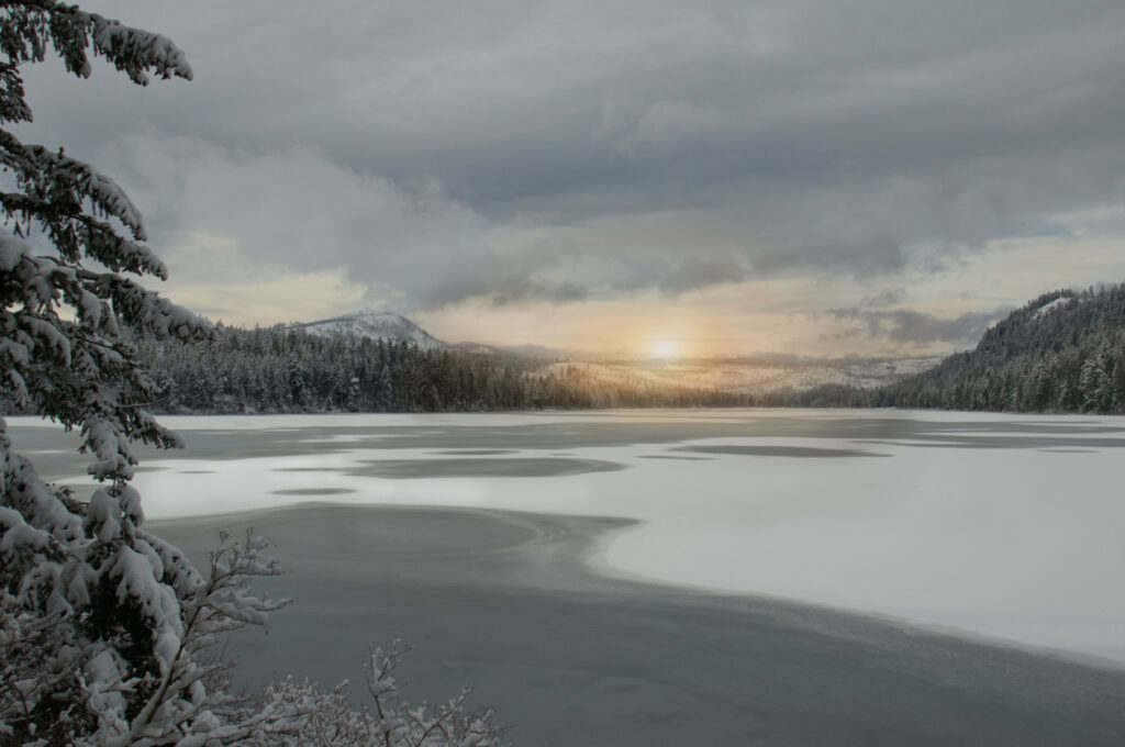 Serene winter landscape with frozen lake snow-covered trees soft sunlight through cloudy sky wallpaper