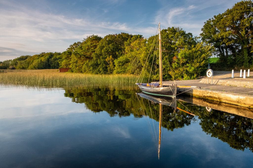 Wooden boat moored at stone pier on calm river with green trees and sky reflections wallpaper