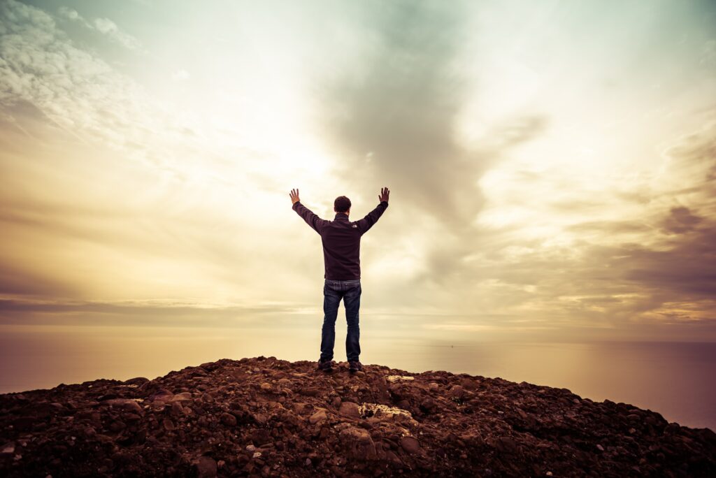 Man triumphantly on rugged mountain peak at sunset with dramatic cloudy sky wallpaper background