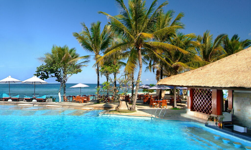 Tropical Bali resort wallpaper with blue pool palm trees thatched-roof hut ocean and clear skies