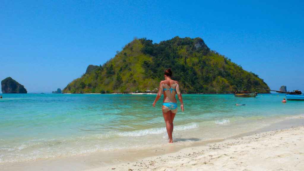 Woman in bikini walking on sandy beach with turquoise water and green island under blue sky Wallpaper