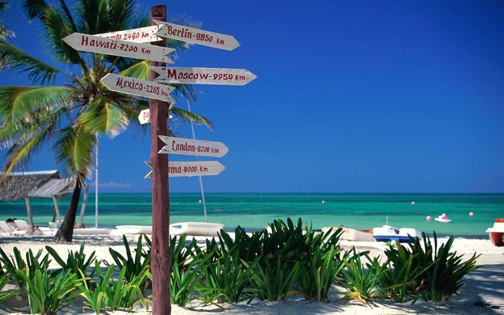 Wooden signpost on white sand beach with turquoise water palm trees and clear blue sky wallpaper