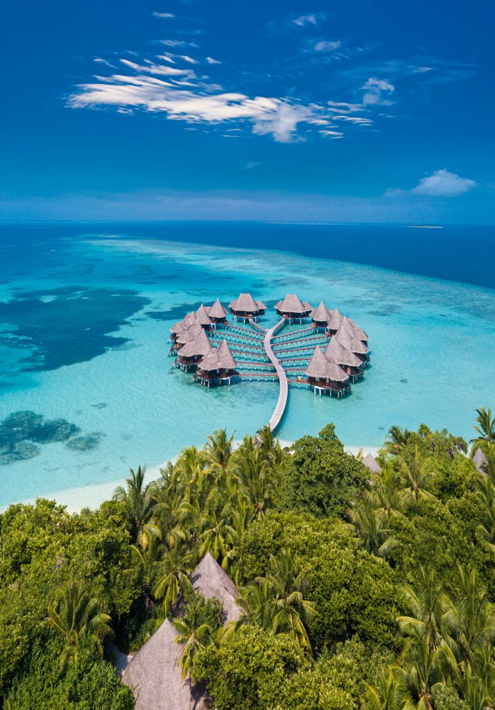 Aerial view of coastal bungalow resort with turquoise waters palm trees and curved pier under blue sky Wallpaper