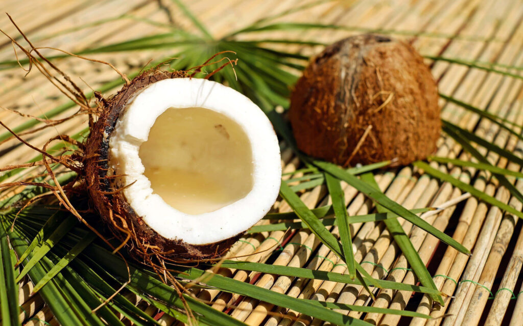 Fresh coconut opened showing white flesh and clear water on palm leaves and bamboo table Wallpaper