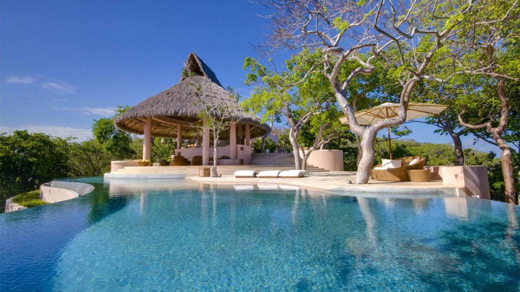Infinity pool with tropical villa thatched-roof cabana sunlit lounge chairs bright blue sky wallpaper