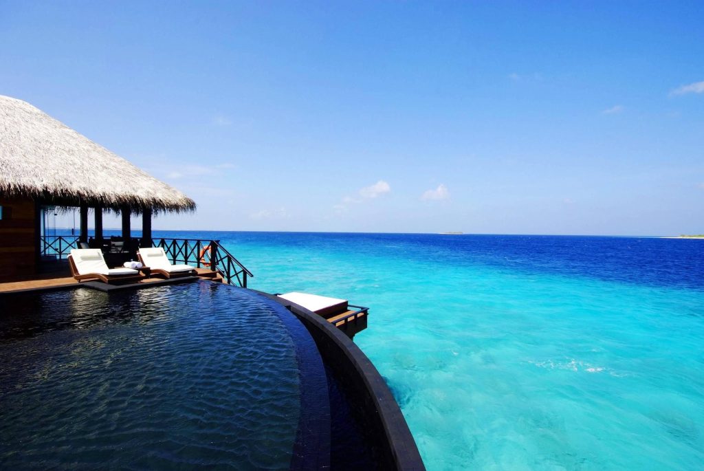 Infinity pool with dark tiles blending into ocean by overwater bungalow under clear skies wallpaper