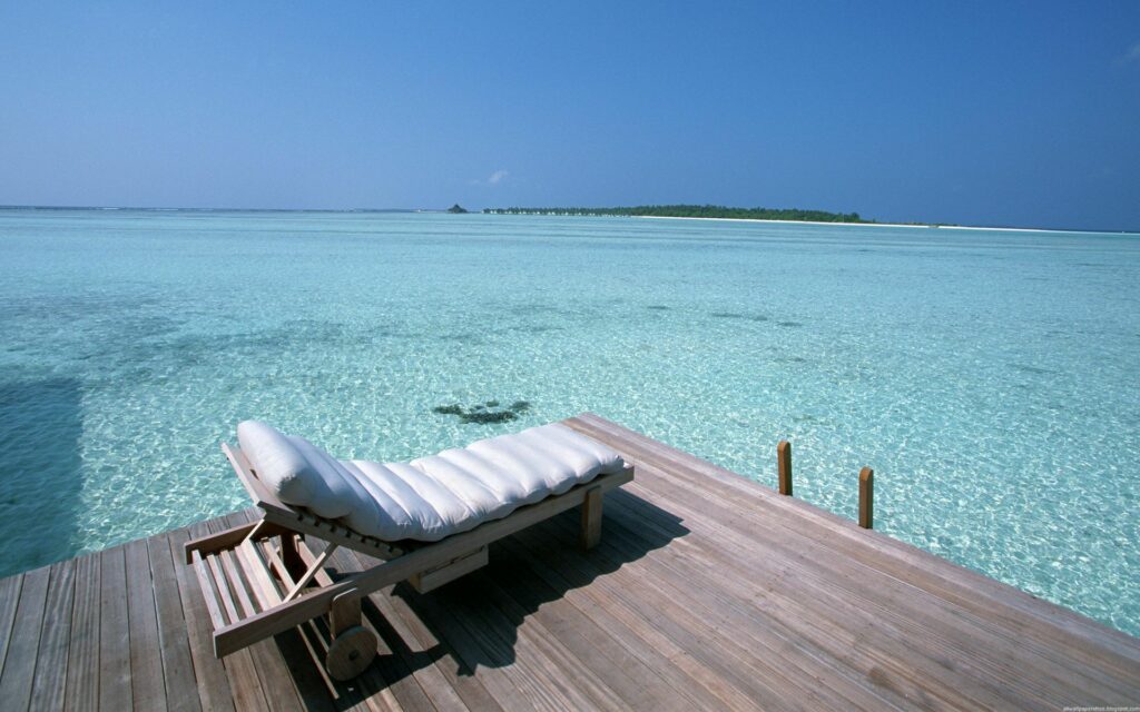 Plush mattress bed on wooden pier over turquoise water under vivid blue sky tropical island view Wallpaper