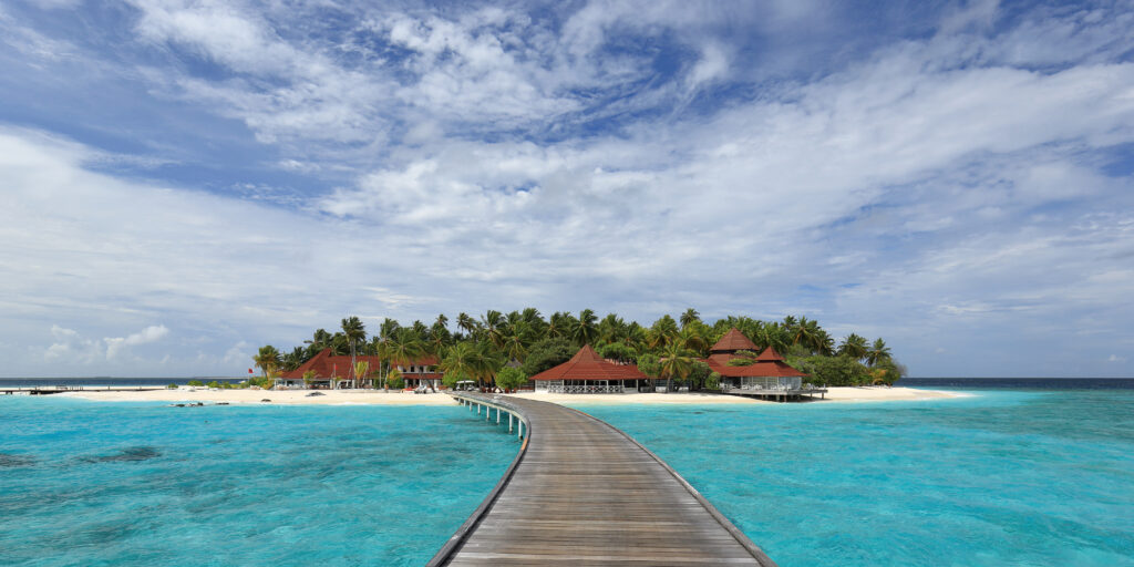 Wooden pier over turquoise tropical waters to island with palm trees red cottages sandy beach Wallpaper
