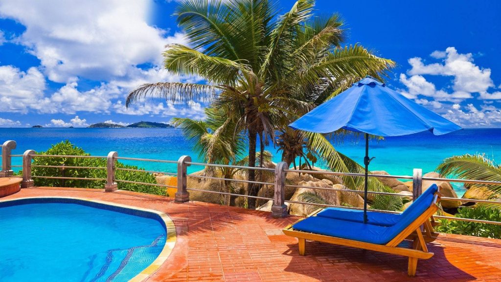 Tropical swimming pool wallpaper with blue parasol lounge chairs ocean palm trees sunny sky