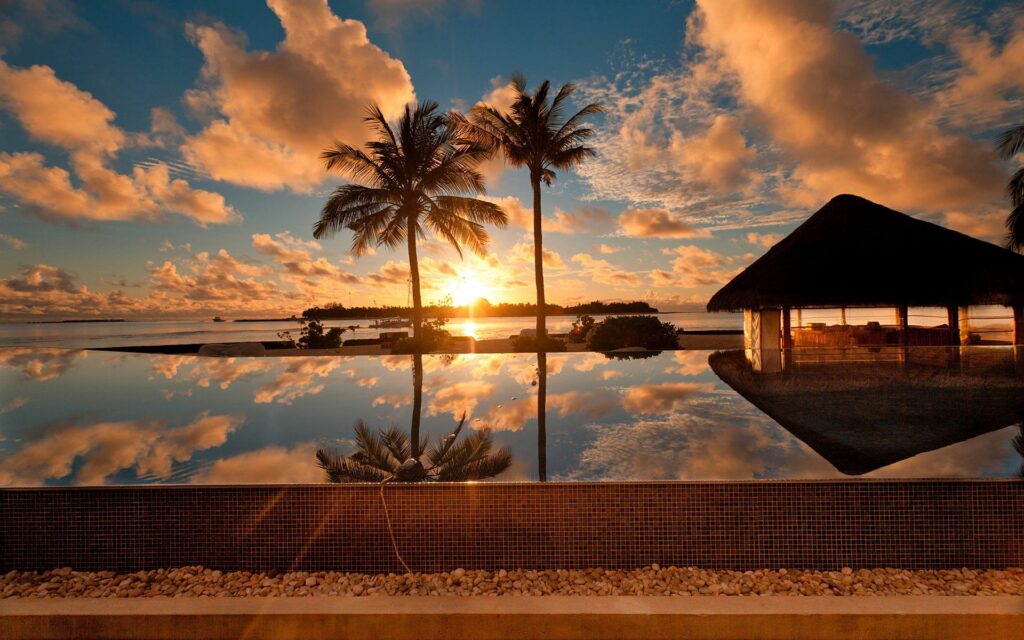 Serene seaside wallpaper with palm trees sunset sky reflective pool and tropical thatched bungalow