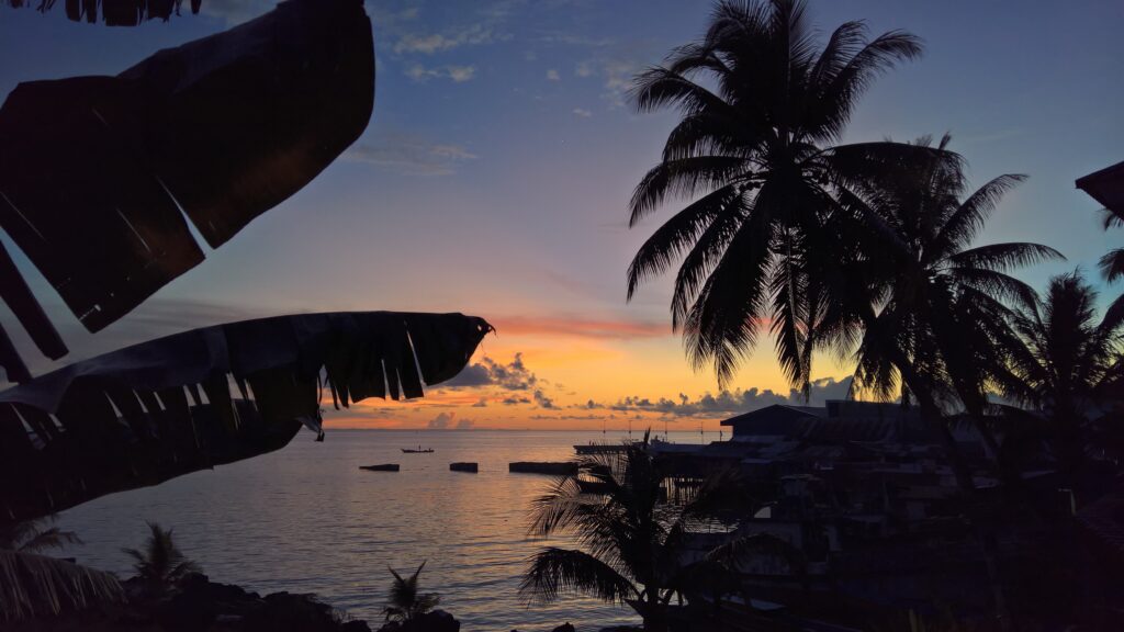 Tropical sunset with orange purple sky palm tree silhouettes calm ocean boats lush foliage wallpaper