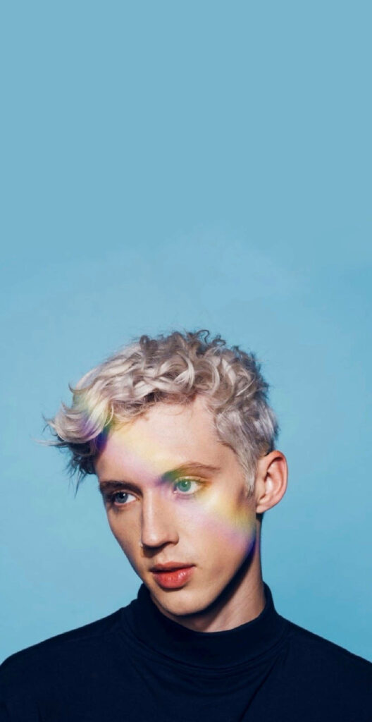 Troye Sivan portrait with rainbow light on face against soft blue background vibrant wallpaper