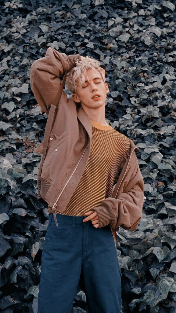 Troye Sivan in stylish brown jacket and striped shirt posing before lush green ivy background wallpaper