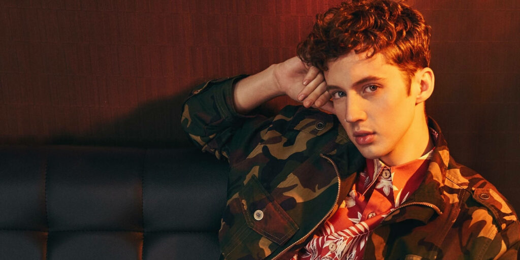 Troye Sivan in camo jacket and bright shirt leaning on sofa warm softly lit background wallpaper