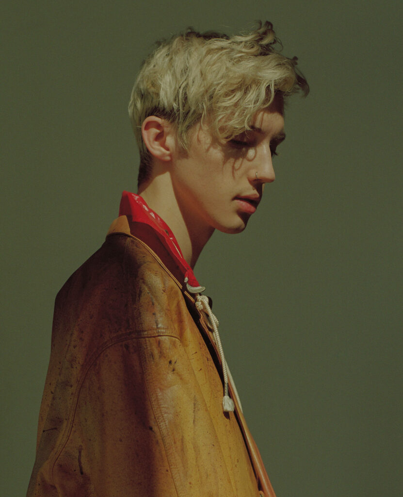 Troye Sivan in vintage jacket and red scarf profile portrait on muted green wallpaper background