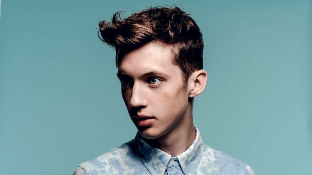 Troye Sivan stylish in light blue patterned shirt on minimalist blue celebrity wallpaper background