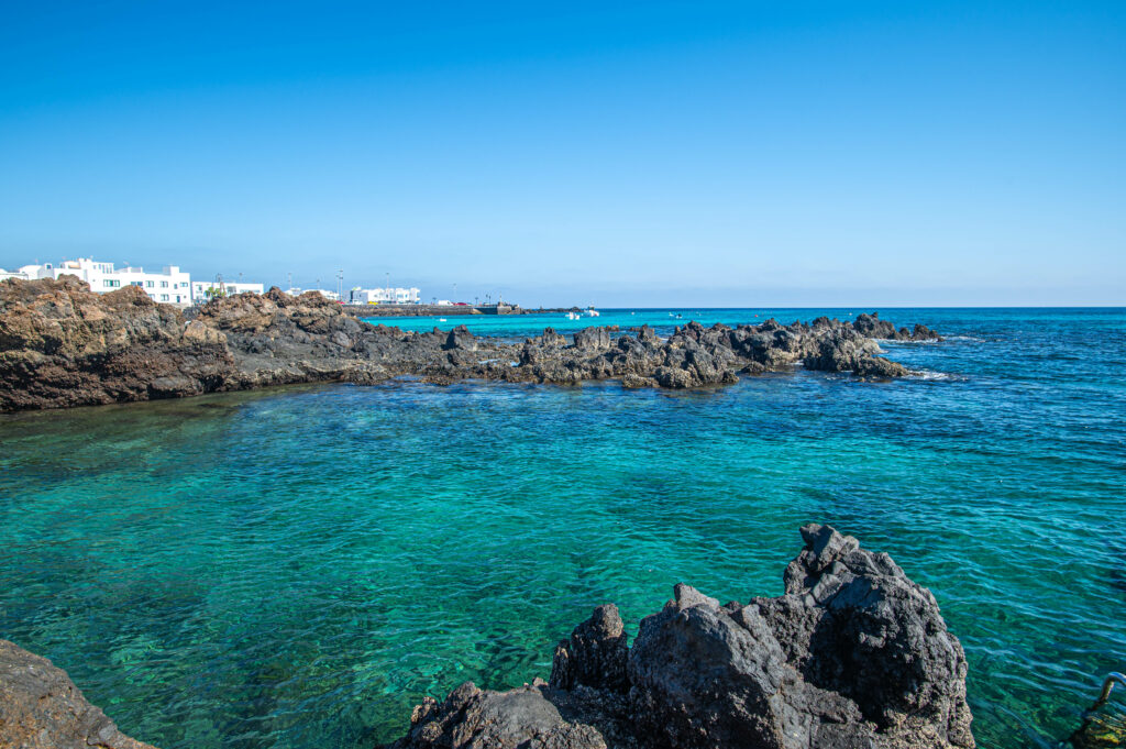Coastal scene with black rocks vibrant reefs turquoise sea white seaside buildings bright horizon Wallpaper