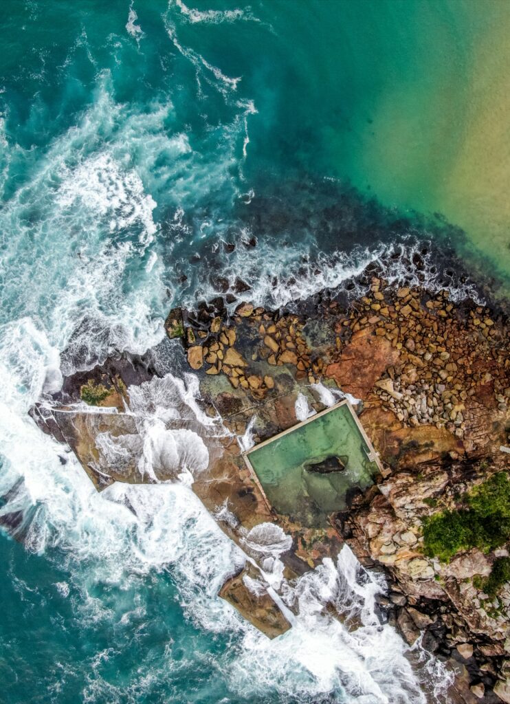 Aerial view of rugged rocky coastline with turquoise sea foamy waves and a seawater pool nestled among stones Wallpaper