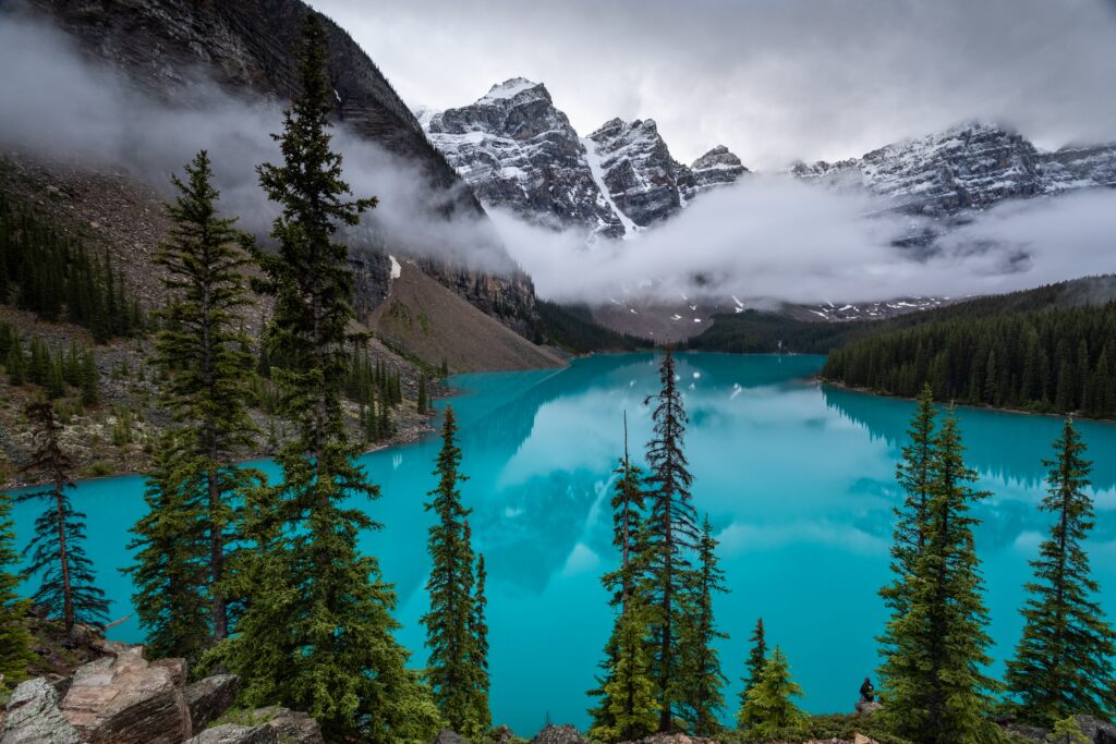 Turquoise lake with evergreen trees snow-capped mountains and low clouds serene landscape wallpaper