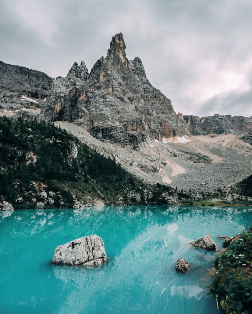 Turquoise lake with rugged rocks lush greenery and mountain peaks reflecting serene sky wallpaper