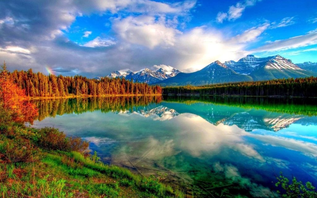 Turquoise lake reflecting snow-capped mountains and pine forests under soft sunlight rainbow Wallpaper