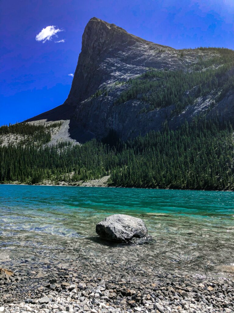 Towering rocky cliff over turquoise lake with clear water smooth pebbles and vivid blue sky wallpaper