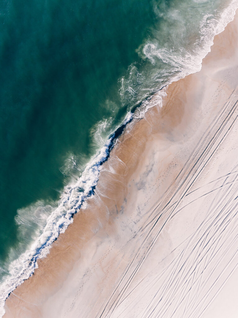 Aerial view of turquoise ocean waves meeting sandy shore with foamy surf and tire tracks wallpaper