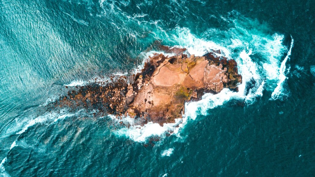 Aerial view of isolated rocky island surrounded by vibrant turquoise ocean waves and surf Wallpaper