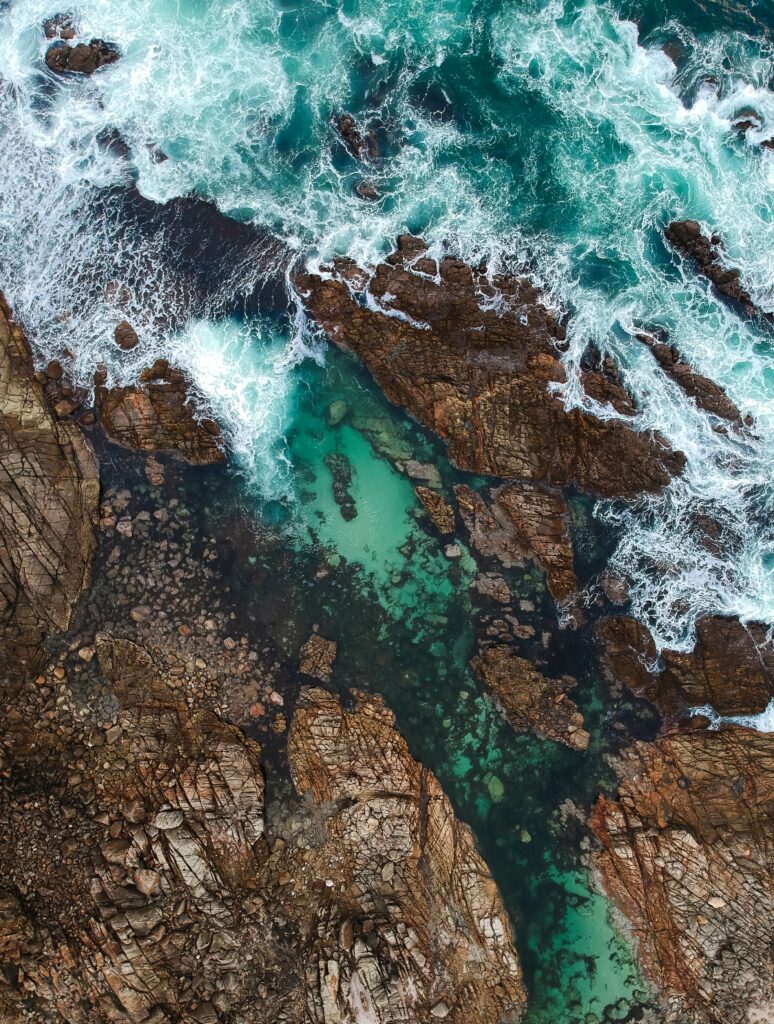 Aerial view of rugged coastal rocks with turquoise waves crashing creating vibrant seascape background Wallpaper